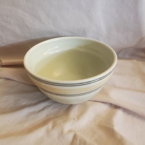 May include: A white ceramic bowl with a striped design in beige and gray. The bowl is empty and sitting on a white surface.