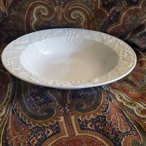 May include: White ceramic bowl with a raised floral design around the rim. The bowl is empty and sitting on a patterned fabric.