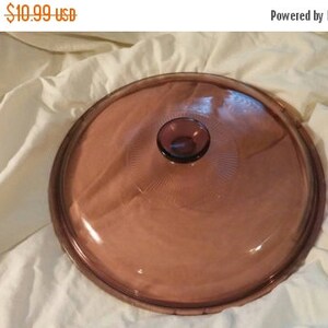 Op de afbeelding: Een ronde, transparante, stoffig roze glazen deksel met een centrale knop. Het deksel heeft een licht verhoogde rand en geëtste concentrische cirkels die uitstralen van de knop. De tekst "Regular Price $10.99 usd" is zichtbaar.