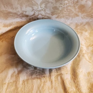 Op de afbeelding: Een lichtblauwe keramische kom met een licht verhoogde rand. De kom is leeg en staat op een wit tafelkleed met een bloemenpatroon.