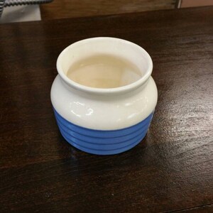 May include: A white ceramic jar with a blue stripe design. The jar is empty and sitting on a dark wooden surface.