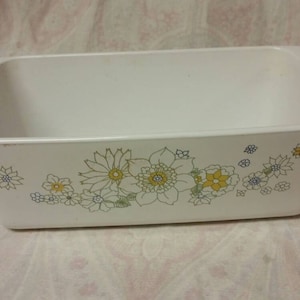 Könnte beinhalten: Weiße Keramik-Backform mit einem Blumendesign in Gelb-, Grün- und Blautönen. Die Form hat eine rechteckige Form mit abgerundeten Ecken und einem leicht erhöhten Rand.