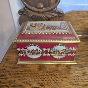 May include: A rectangular, burgundy-colored trinket box with a gold-colored frame. The lid features a detailed illustration of a horse-drawn carriage. The sides display oval illustrations of horse-drawn carriages and winter scenes. A decorative item.