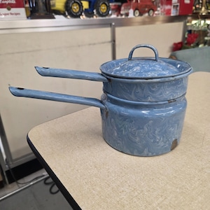 Puede incluir: Una antigua olla de doble caldera de esmalte azul con tapa. La olla tiene dos asas largas y un patrón veteado. La olla está sobre una mesa de color claro.