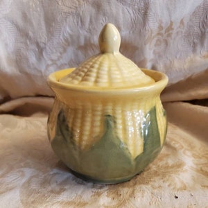 Peut inclure: Un pot à sucre en céramique jaune et vert avec un couvercle en forme d'épi de maïs. Le pot a une surface texturée qui ressemble à des grains de maïs.