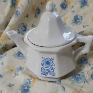 Puede incluir: Tetera de cerámica blanca con un diseño floral azul. La tetera tiene una tapa y un asa. La tetera está sentada sobre un mantel floral blanco y amarillo.