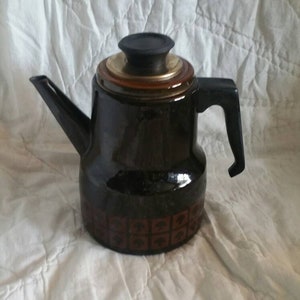 May include: A dark brown ceramic teapot with a black handle and spout. The teapot has a decorative pattern around the base and a black lid. The top of the teapot has a gold-coloured band.