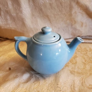 May include: A light blue ceramic teapot with a rounded body and a lid with a small spout. The teapot is sitting on a white fabric surface.