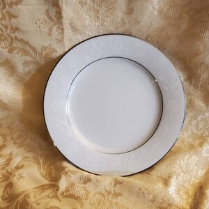 Puede incluir: Un plato de cena de cerámica blanca con un borde dorado y un delicado patrón floral. El plato está sobre un fondo de tela dorada.