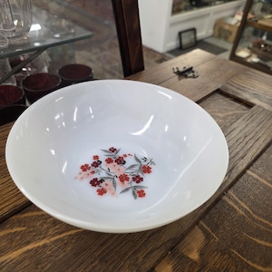 May include: A white, round bowl featuring a floral pattern in the centre. The flowers are red and pink with grey leaves. The bowl rests on a wooden surface.