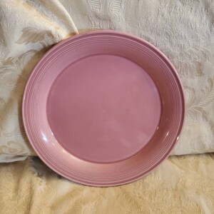 Op de afbeelding: Een ronde, roze plaat met een geribbelde rand. De plaat is effen van kleur en lijkt gemaakt van keramiek of een vergelijkbaar materiaal. De plaat ligt op een crèmekleurige stof met een patroon.