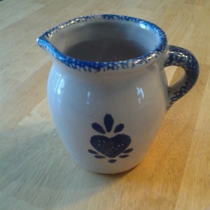 Puede incluir: Un peque&ntilde;o creamer de cer&aacute;mica blanca con un dise&ntilde;o floral azul y blanco. El creamer tiene un asa y un pico.