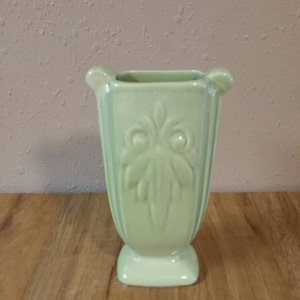 Puede incluir: Un jarrón de cerámica verde pálido con una base cuadrada y un diseño acanalado. El jarrón tiene un patrón floral decorativo.