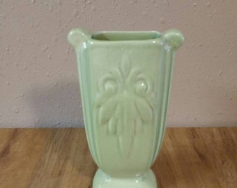 May include: A pale green ceramic vase with a square base and a fluted design. The vase has a decorative floral pattern.
