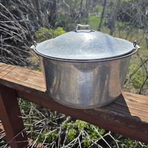 May include: A vintage, aluminum cooking pot with a lid. The pot is silver and has a handle on the lid. The pot is sitting on a wooden surface, with a natural background of trees and foliage.