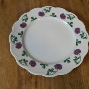 May include: White ceramic dinner plate with a scalloped edge and a decorative border of green leaves and purple clover flowers.
