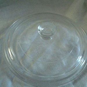 Op de afbeelding: Een heldere glazen deksel met een ronde knopgreep. De deksel is cirkelvormig met een iets verhoogde rand en is ontworpen om op een pot of pan te passen. Het glas is transparant, waardoor de inhoud zichtbaar is.