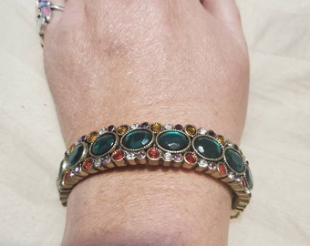 May include: A gold-toned bracelet with a row of oval-shaped green gemstones surrounded by smaller clear and colored gemstones.
