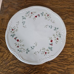 May include: A round, white ceramic plate with a scalloped edge. The plate is decorated with a holly and berry pattern in green, red, and white. The plate is sitting on a wooden surface.