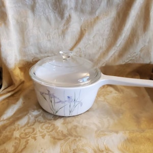 May include: White ceramic saucepan with a glass lid and a floral design featuring purple irises. The handle is white.