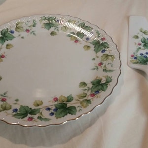 May include: A white ceramic cake plate with a green and floral design. The plate has a scalloped edge and a gold trim. A matching cake server is also included.
