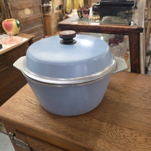 May include: A light blue Dutch oven with a matching lid and a dark brown knob handle. The pot features a silver-toned metal rim and two integrated side handles. The exterior has a smooth, matte finish, and the overall design is circular.
