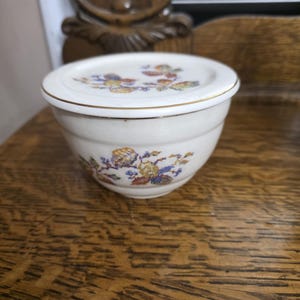 May include: A white ceramic butter dish with a lid. The dish and lid are decorated with a floral pattern in shades of blue, yellow, and red. The rim of the lid has a gold trim. The dish is sitting on a wooden surface.