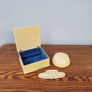 Può includere: Un set di tre articoli da toeletta vintage di colore giallo pallido. Il set comprende una piccola scatola con un interno in velluto blu, un barattolo rotondo con un coperchio e un piatto rettangolare con un manico rialzato.