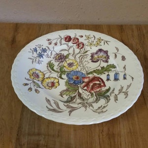 May include: A white ceramic dinner plate with a floral design. The flowers are in shades of red, yellow, blue, and purple. The plate has a raised rim and a scalloped edge.