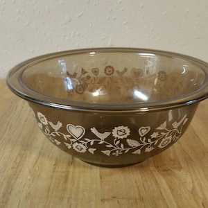 May include: A brown glass mixing bowl with a white floral design featuring birds and hearts. The bowl has a clear glass rim.