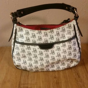 May include: A white and black Dooney & Bourke handbag with a black leather strap and red lining. The bag is covered in a repeating pattern of the Dooney & Bourke logo.