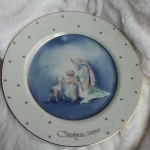 Pu&ograve; includere: Un piatto in ceramica bianca con bordo dorato e un'immagine blu e bianca di una donna e due bambini. Il piatto ha un'iscrizione dorata che recita "Christmas 1980".