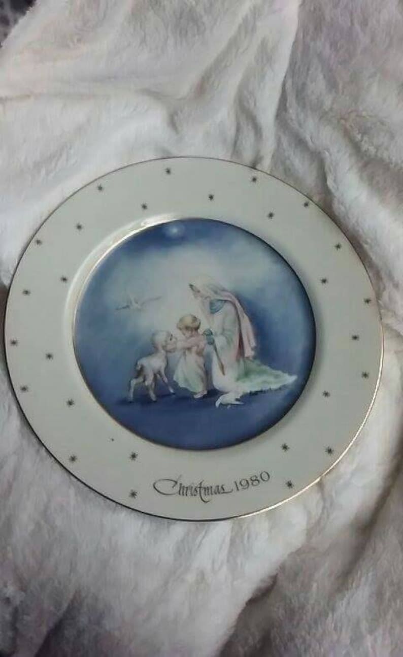 Pu&ograve; includere: Un piatto in ceramica bianca con bordo dorato e un'immagine blu e bianca di una donna e due bambini. Il piatto ha un'iscrizione dorata che recita "Christmas 1980".