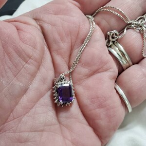 Może przedstawiać: Srebrny łańcuszek z naszyjnikiem, na którym znajduje się duży, prostokątny, fioletowy ametyst otoczony mniejszymi, przezroczystymi kamieniami szlachetnymi.