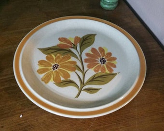 May include: A white ceramic dinner plate with a brown rim and a floral design featuring orange and yellow flowers with green leaves.