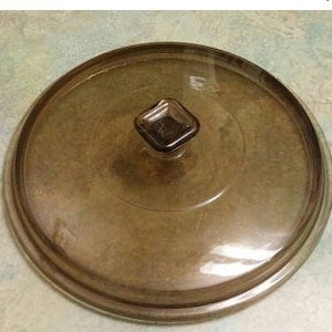 Puede incluir: Una tapa de vidrio transparente con un borde marrón y una perilla cuadrada en la parte superior. La tapa está diseñada para una olla o sartén.
