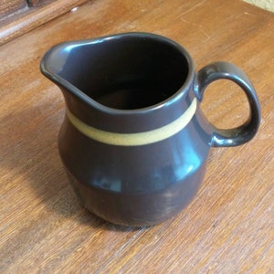 Puede incluir: Una jarra de crema de cerámica marrón con una sola franja marrón alrededor del centro. La jarra tiene un cuerpo redondeado y un asa.