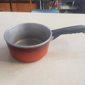 May include: A small orange and black saucepan with a black handle. The saucepan is made of aluminum and has a non-stick coating.