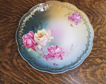 May include: A vintage porcelain plate with a scalloped edge and a floral design. The plate features pink, yellow, and white roses on a blue and green background.