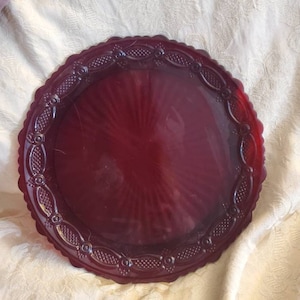 May include: A circular, deep red glass plate with a decorative, scalloped edge and an ornate border. The plate has a textured, radial pattern. The border features a repeating design of oval shapes and small dots.