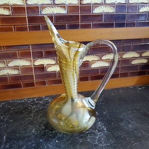 Może przedstawiać: Żółty dzbanek szklany z białym wzorem w postaci spirali. Dzbanek ma okrągłe dno i długą, cienką szyjkę z dzióbkiem. Ucho jest ze szkła przezroczystego.