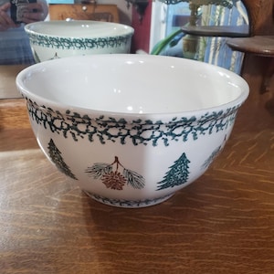 Puede incluir: Tazón de mezcla de cerámica blanca con un diseño de árbol de pino verde y marrón. El tazón tiene un borde ancho y un fondo redondeado.