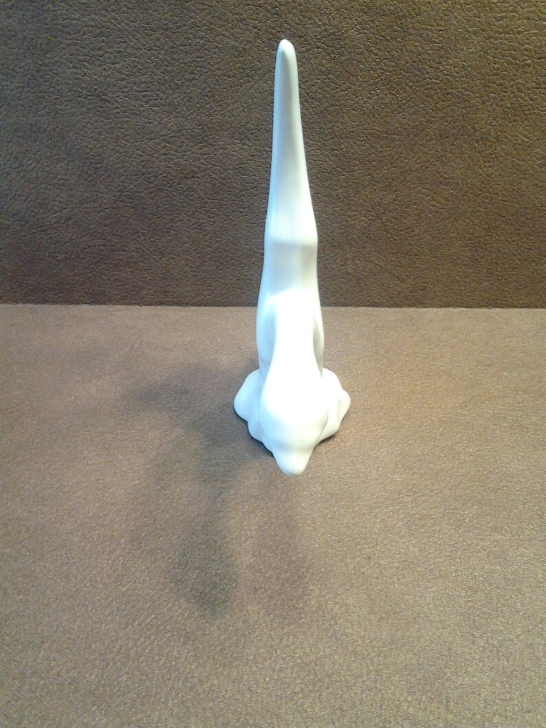 May include: A white ceramic bunny figurine in a yoga pose, with its head touching the ground and its body upright. The bunny is on a brown surface. The figurine has a smooth, glossy finish and a minimalist design.