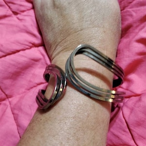 May include: A silver-toned metal bracelet with a unique, curved design. The bracelet is made of multiple bands that are intertwined and create a geometric pattern.