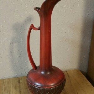 Può includere: Un vaso in ceramica rosso con un disegno marrone intorno alla base. Il vaso ha un'ampia apertura e un manico.