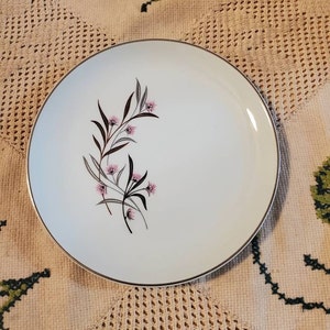 Könnte beinhalten: Weißer Keramik-Dinnerteller mit silbernem Rand und einem Blumendesign mit rosa Blüten und dunkelgrünen Blättern.