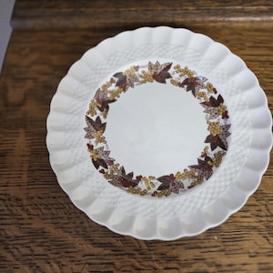 May include: A white, scalloped-edge plate with a woven texture and a central design of autumn leaves and berries in shades of brown, gold, and red. The plate is set against a wooden surface.