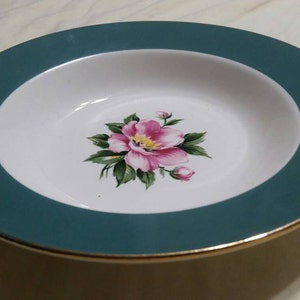 May include: A white soup bowl with a teal rim and a gold trim. The bowl features a pink flower with green leaves.