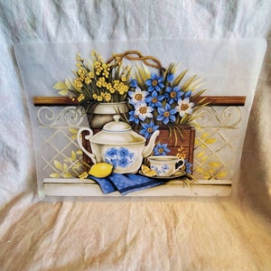 May include: A white teapot with blue floral designs and a matching teacup and saucer. The teapot is sitting on a blue and white patterned napkin with a lemon beside it. A bouquet of yellow and blue flowers is in a brown pot behind the teapot.