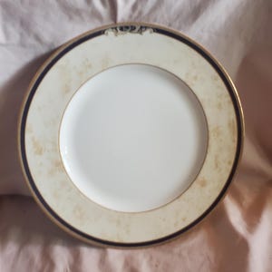Puede incluir: Un plato de cena de cerámica blanca con un borde dorado y negro. El plato tiene un diseño floral sutil en el centro.